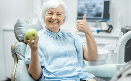senior woman in dental chair holding green apple and giving thumbs up 